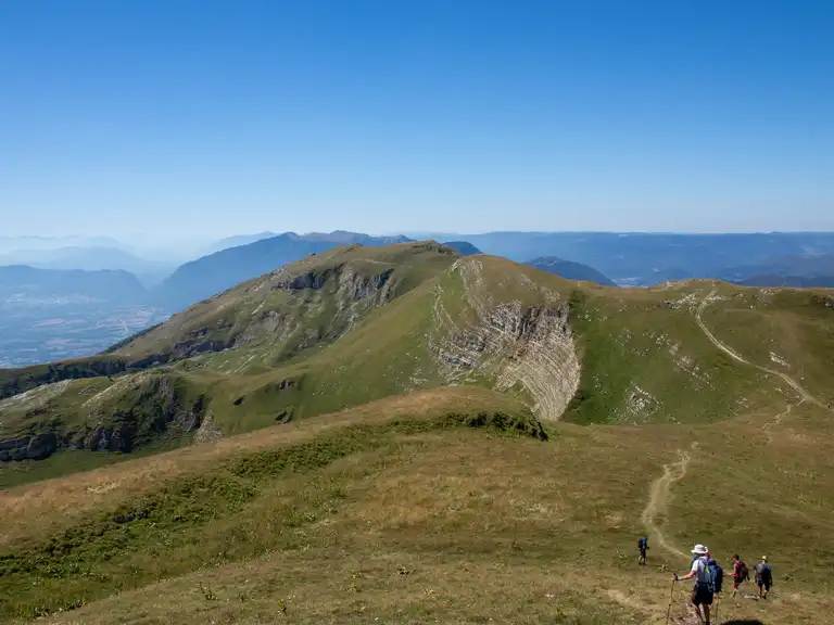 Randonneurs marchant sur un sentier de montagne verdoyant avec une vue panoramique sur les collines et montagnes au loin, sous un ciel bleu clair.