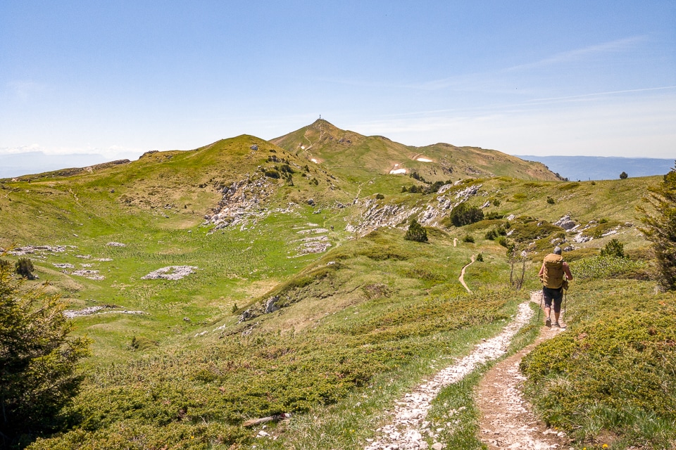 Randonnée vers le Crêt de la Neige avec un sentier traversant des collines verdoyantes, un randonneur avec un sac à dos et des vues panoramiques sur les montagnes