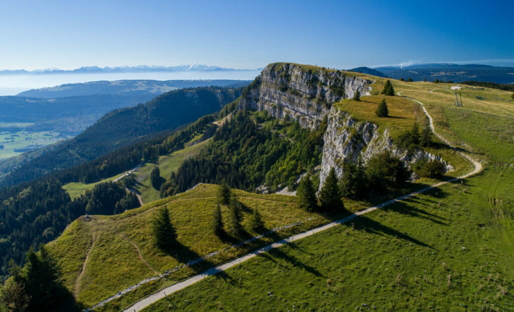 Vue aérienne du sommet du Mont d'Or dans le Pays de Gex, montrant un chemin pour vélo, sinueux à travers des collines verdoyantes avec des falaises abruptes et des forêts denses en arrière-plan, sous un ciel bleu clair.