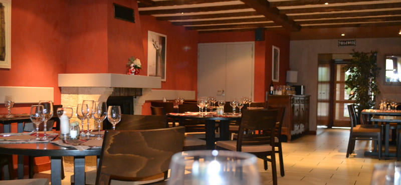 Salle à manger d'un restaurant dans le Pays de Gex avec des murs rouge foncé, des tables en bois bien dressées, des verres à vin et une cheminée en arrière-plan, créant une ambiance chaleureuse et élégante