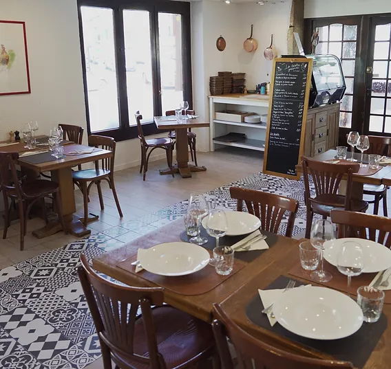 Intérieur d'un restaurant dans le Pays de Gex avec des tables en bois bien dressées, des assiettes blanches et des verres à vin, créant une ambiance accueillante et chaleureuse, avec un tableau noir affichant le menu du jour en arrière-plan