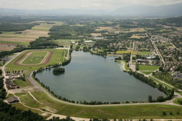 Vue aérienne du lac de Divonne-les-Bains dans le Pays de Gex, entouré de verdure, avec des installations sportives et des habitations visibles en arrière-plan, permettant de faire du vélo.