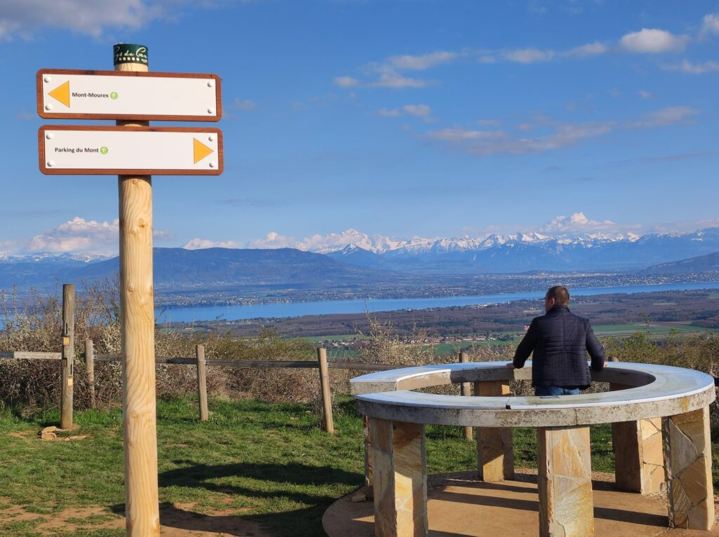 Vue panoramique depuis le Mont Mourex dans le Pays de Gex, avec des panneaux de signalisation indiquant les directions et un homme contemplant le paysage, incluant le lac Léman et les montagnes en arrière-plan.